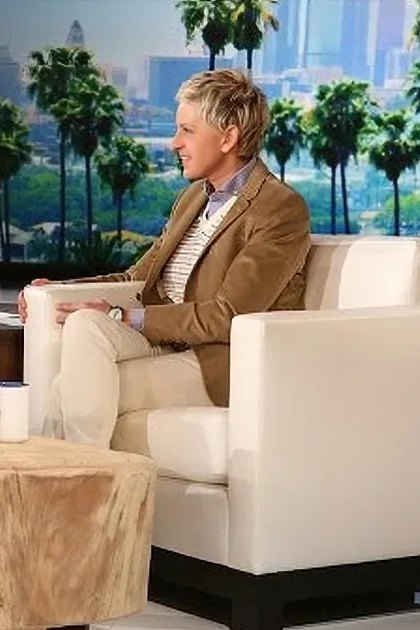 Talk show set with two couches, tree bark-covered coffee tables, white armchairs and sofa, small green plant, mugs on the table; large windows in background showing cityscape and palm trees. Daytime setting with natural light.