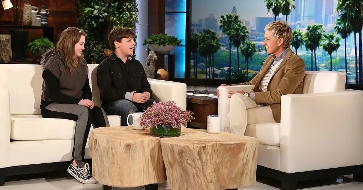 Talk show set with two couches, tree bark-covered coffee tables, white armchairs and sofa, small green plant, mugs on the table; large windows in background showing cityscape and palm trees. Daytime setting with natural light.