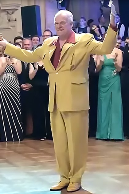 An adult male singer in a bright yellow suit dancing with an older adult woman in a floral dress at an elegant indoor event.