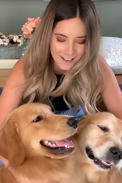 Young adult female wearing a sleeveless top and jeans, holding two golden retriever dogs indoors with wooden table and cushioned bench visible.