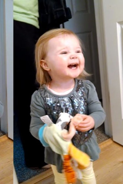 A toddler in an indoor setting, wearing a dark green long-sleeved shirt with sequins and light yellow pants. The background includes white doors and a dark blue carpeted area.