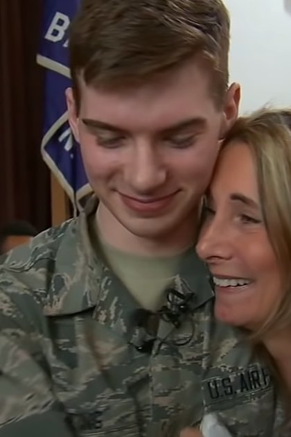 A young male soldier in U.S. Air Force uniform embraces a middle-aged woman with shoulder-length blonde hair, standing indoors within what appears to be a school setting during an event or assembly related to military appreciation.