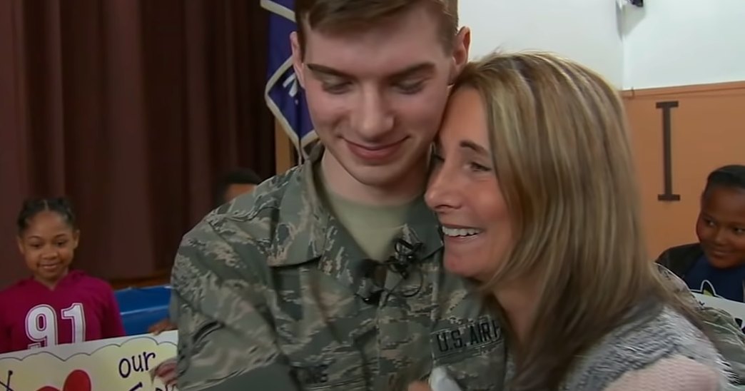 A young male soldier in U.S. Air Force uniform embraces a middle-aged woman with shoulder-length blonde hair, standing indoors within what appears to be a school setting during an event or assembly related to military appreciation.