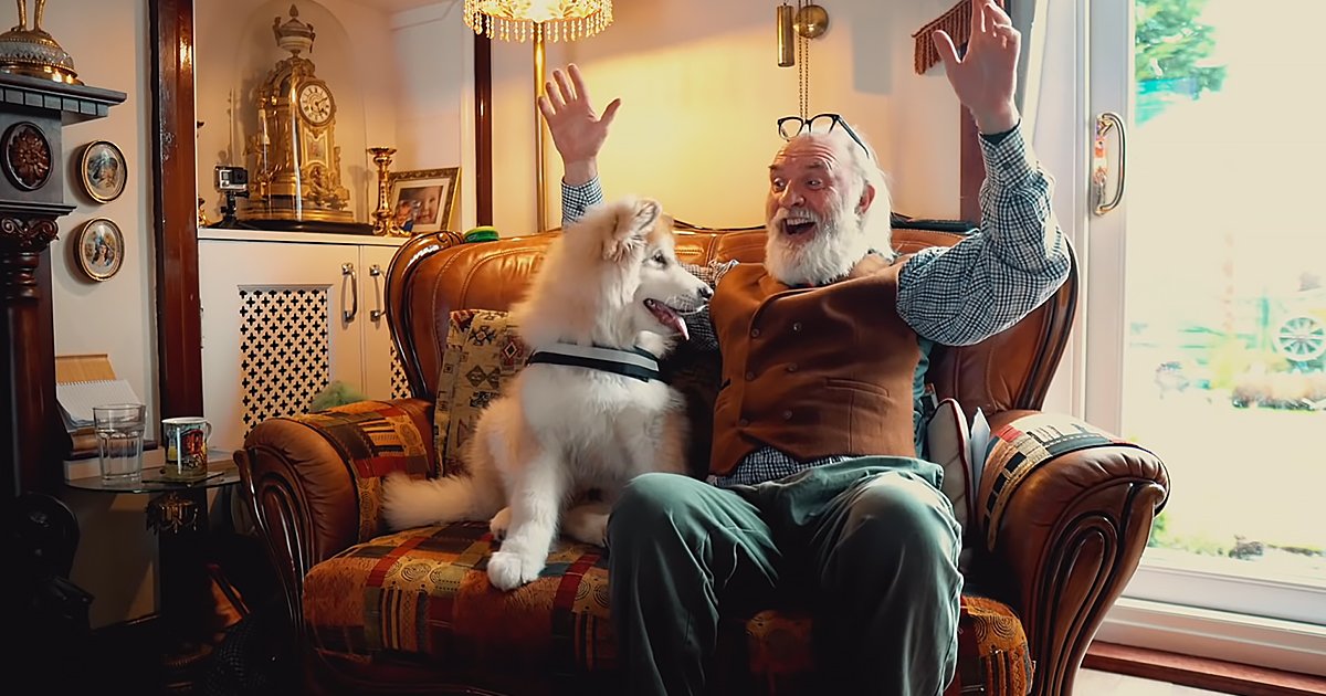 An adult male wearing a checkered shirt under a brown vest sits with an adult Samoyed dog in a cozy living room filled with warm lighting and spring-greenery visible through the window.