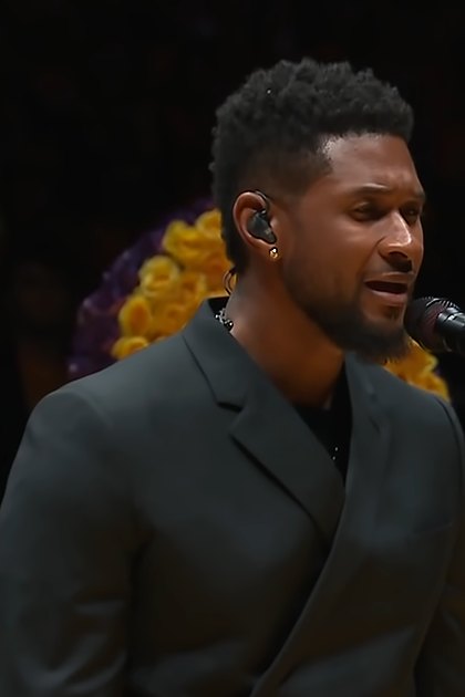 A young adult male performer in a dark gray suit jacket, possibly at an indoor memorial service. Large floral tributes with the number '24' are prominently displayed in the background.