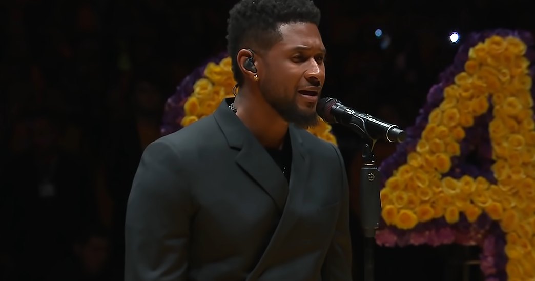 A young adult male performer in a dark gray suit jacket, possibly at an indoor memorial service. Large floral tributes with the number '24' are prominently displayed in the background.