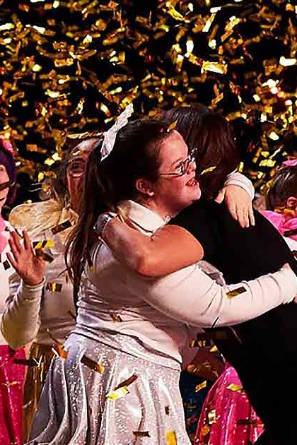 A group celebration indoors with festive attire and confetti falling throughout the scene; evening or nighttime lighting illuminates a gathering of young adults in various colors.