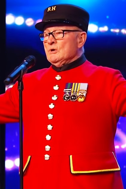 A male performer in a red military-style jacket adorned with medals, performing at an indoor stage event under bright lighting and colorful effects.