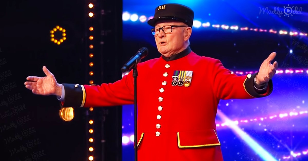 A male performer in a red military-style jacket adorned with medals, performing at an indoor stage event under bright lighting and colorful effects.