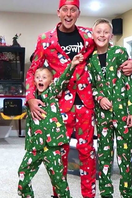 Adult Female carrying clothes, wearing light gray top and black pants with rips; Adult Male in red Santa suit jacket over t-shirt and green pajama-style pants; Child in green Santa-themed pajama set standing near adult male; Teen/Young Adult Male in similar attire to the adult male. Scene includes dark brown couch, arcade machine, television showing colorful display, plastic storage bins, baby swing with toys attached.