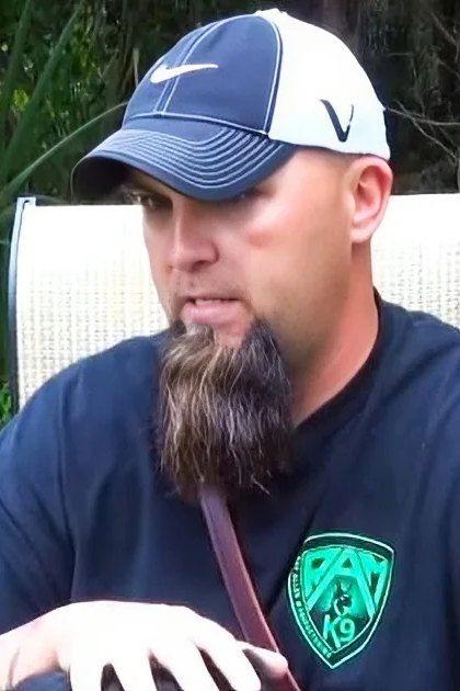 Adult male in a relaxed casual t-shirt with 'RAT 19' logo and white Nike cap, sitting outdoors on green grassy terrain with white woven chair visible behind him.