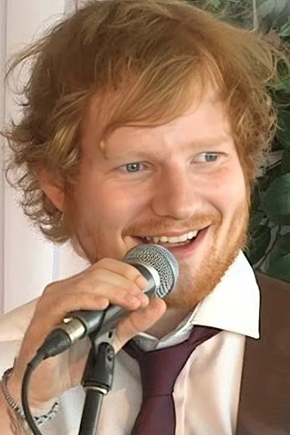 A young adult male with reddish-brown hair and beard sings into a microphone at an indoor event while wearing a white dress shirt, purple tie, and brown suspenders.