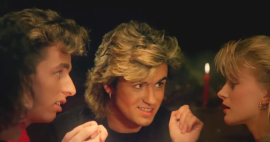 A young adult male stands in a dimly lit indoor setting with two other individuals flanking him; one is a female on his left, another on the right.