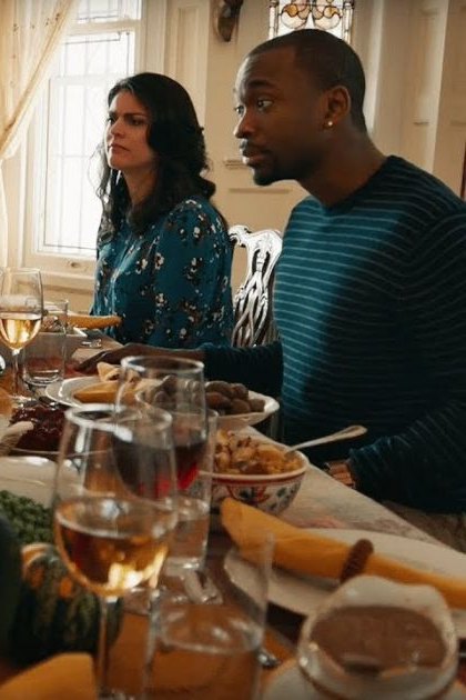 A family gathers around a dining table set for a Thanksgiving meal in an indoor setting decorated with natural light and autumnal motifs.