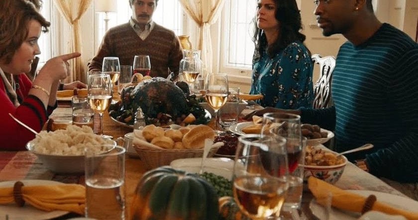 A family gathers around a dining table set for a Thanksgiving meal in an indoor setting decorated with natural light and autumnal motifs.