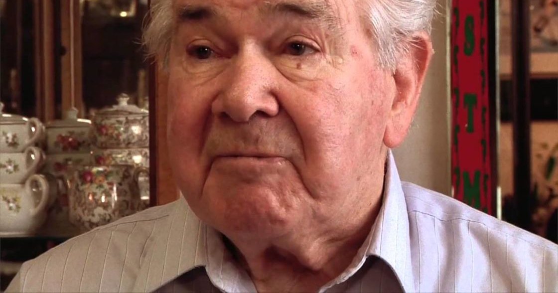 An older adult male wearing a light gray, vertically striped collared shirt in an indoor setting with teapots and decorative items.