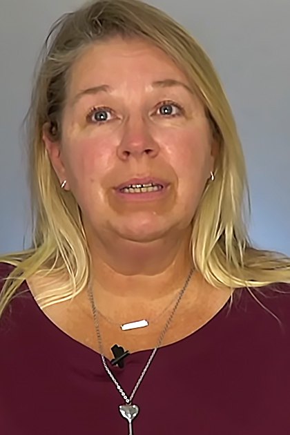 Middle-aged adult female wearing a maroon, long-sleeved, scoop-neck, relaxed-fit top with a small black microphone necklace and earrings against a neutral gray or light blue wall indoors.