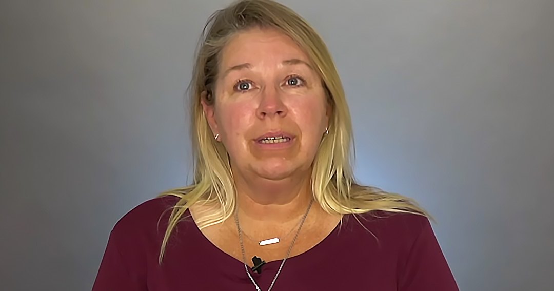 Middle-aged adult female wearing a maroon, long-sleeved, scoop-neck, relaxed-fit top with a small black microphone necklace and earrings against a neutral gray or light blue wall indoors.