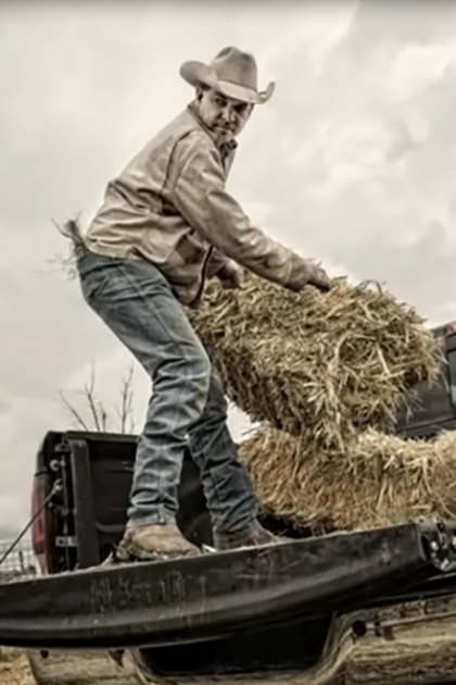 A middle-aged man in a light-colored shirt and blue jeans loads hay into a black pickup truck on an overcast day in an open field with dry grass, wire fence, and trees.