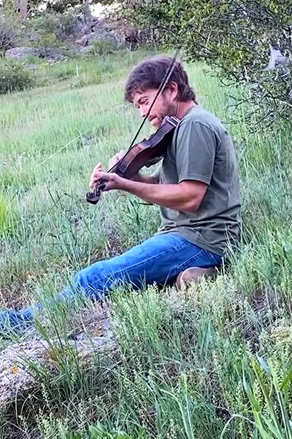 An adult male playing a violin in an outdoor natural setting with a red fox nearby, lush greenery and hills visible.