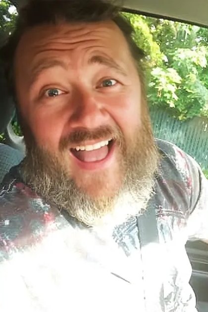 A middle-aged adult male wearing a patterned shirt with long sleeves sits outdoors amidst greenery, suggesting spring or summer under natural daylight.
