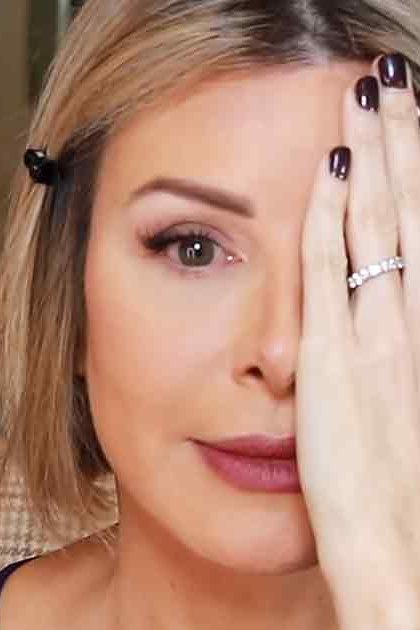 A middle-aged adult female wearing a dark-colored top, ring finger adorned with a visible ring and dark nail polish, in an indoor residential setting with a staircase, framed pictures, and decorative branches.