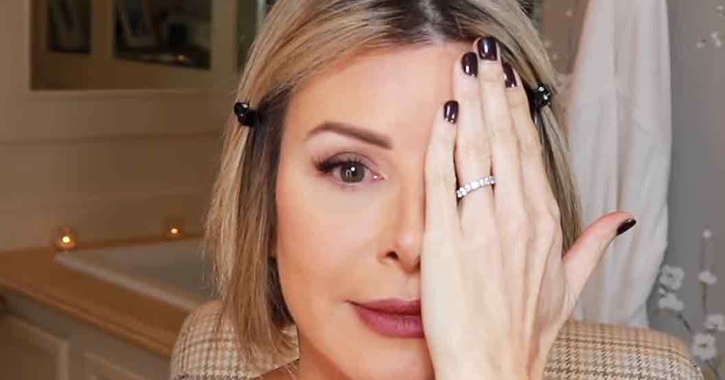A middle-aged adult female wearing a dark-colored top, ring finger adorned with a visible ring and dark nail polish, in an indoor residential setting with a staircase, framed pictures, and decorative branches.