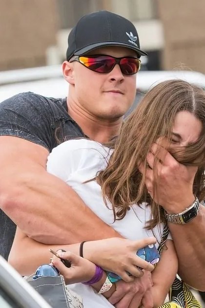 A muscular man in a cap and sunglasses carries a woman covering her face through a parking lot.