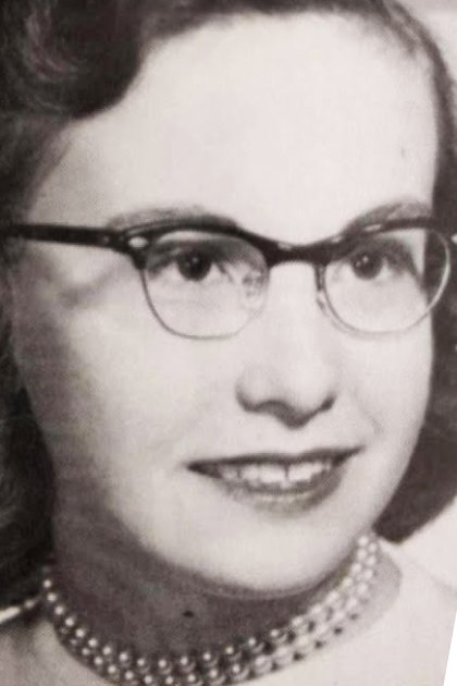 Middle-aged adult female wearing pearl necklace, glasses with thick frames, light-colored garment; purple handbag with geometric pattern in foreground indoors.
