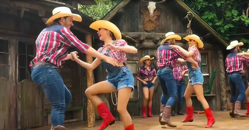 A group dancing in a rustic setting, likely at an amusement park or themed attraction.