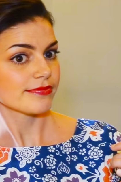 A young adult female wearing a floral dress with blue background and orange, purple, and white flowers indoors.