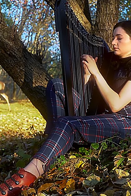 A young adult female plays a black harp in an autumn forest setting with long shadows and fallen leaves. A white-tailed deer is nearby, blending into the natural environment.