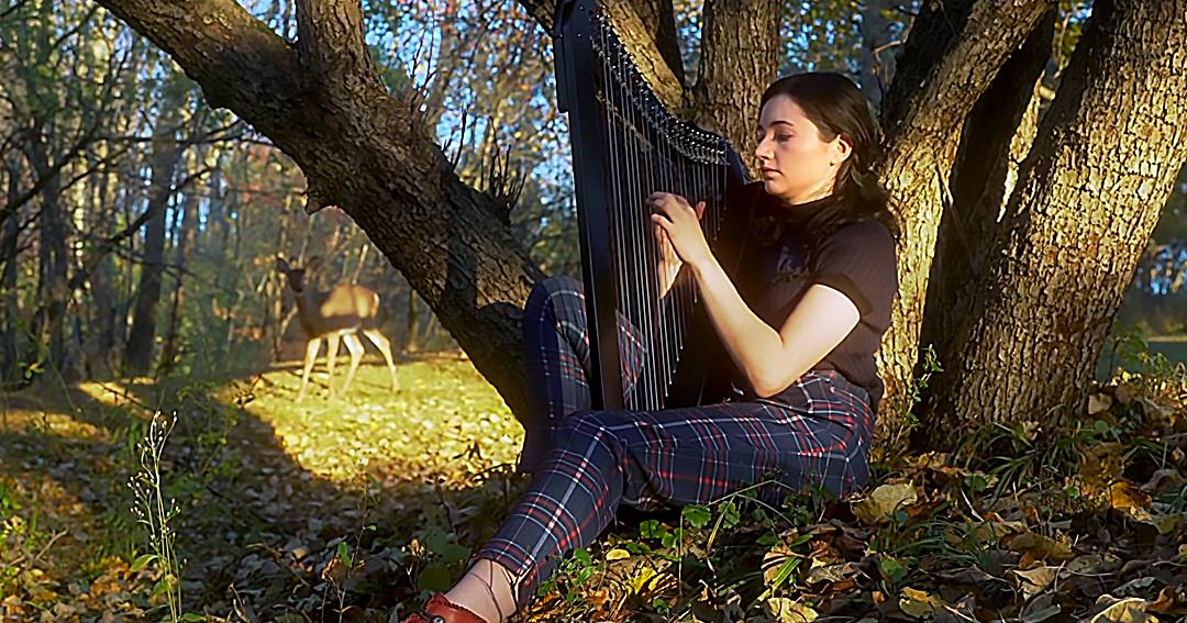 A young adult female plays a black harp in an autumn forest setting with long shadows and fallen leaves. A white-tailed deer is nearby, blending into the natural environment.